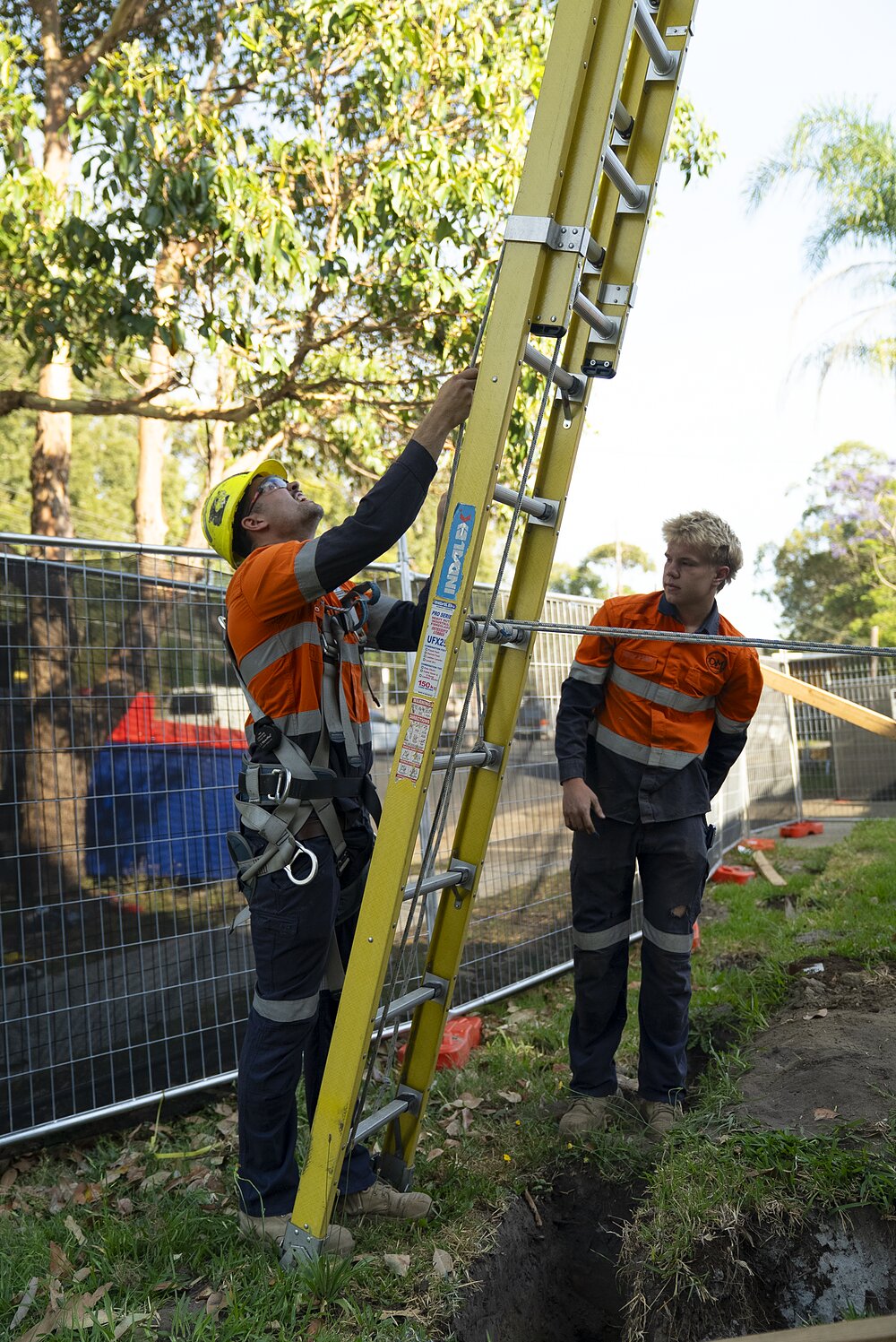 Two DM Electrical electricians in hi-vis and hard hats on a Sydney job site, one on a ladder beside an open service trench